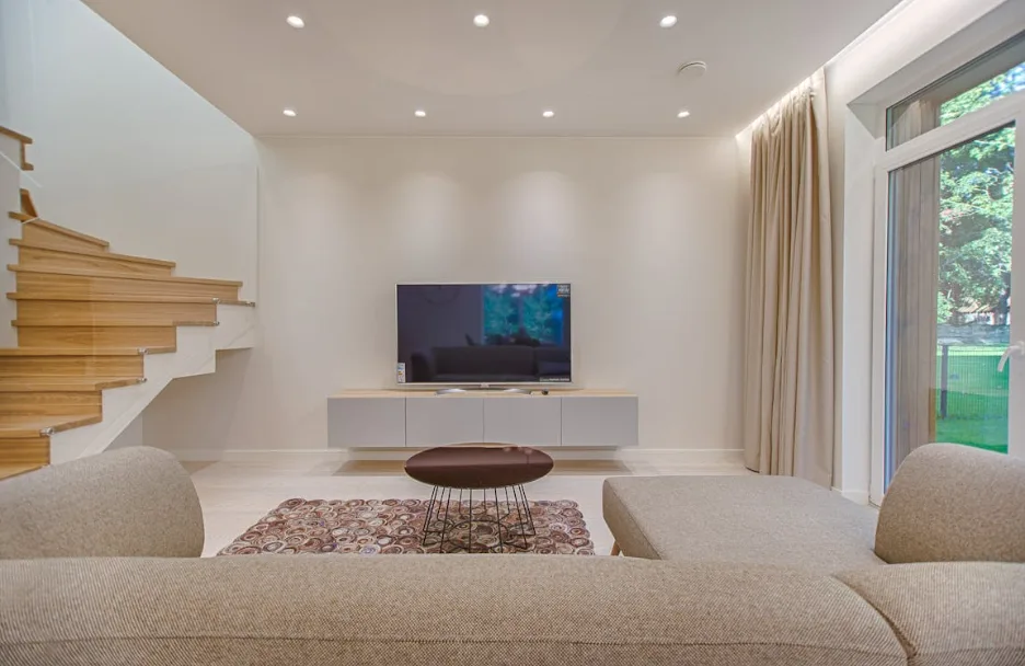Modern living room with beige sofa, flat-screen TV, wooden staircase, large window with curtains, and minimalist décor.
